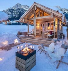 a small wooden cabin in the snow with fire pit and chairs around it at night