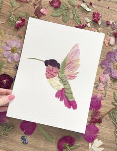 a person holding up a piece of paper with flowers around it and a hummingbird painted on it