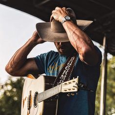 a man with a cowboy hat holding his guitar up to his head while wearing a blue shirt and jeans