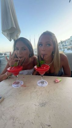 two beautiful women sitting at a table with drinks in front of their faces and making the peace sign