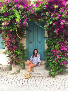 a woman sitting in front of a blue door surrounded by purple flowers