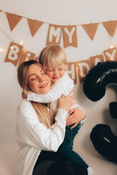 a woman holding a child in front of a happy birthday banner with balloons and bunting