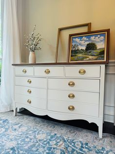 a white dresser with gold handles and drawers in front of a painting on the wall