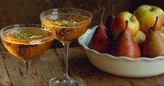 two glasses filled with wine and pears next to a bowl of fruit on a table