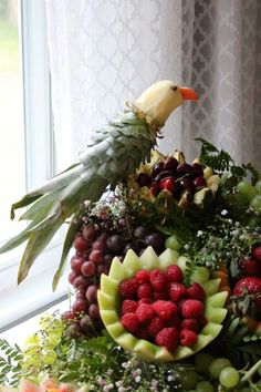 an arrangement of fruits and vegetables in front of a window with a bird on top