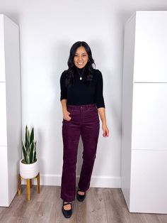 a woman in black shirt and purple pants standing next to a white wall with potted plant