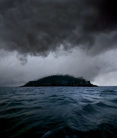 an island in the middle of the ocean under a dark sky with storm clouds above it