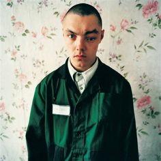 a young man wearing a green jacket and white shirt with flowers on the wall behind him