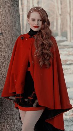 a woman with long hair wearing a red cape and black dress standing next to a tree