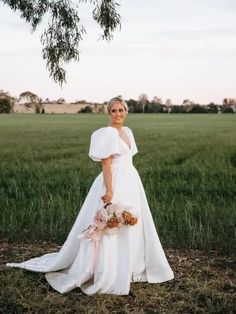 a woman in a white wedding dress standing under a tree with her hands on her hips