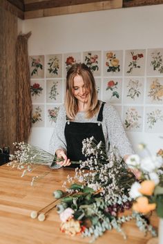 a woman standing in front of a wooden table with flowers and greenery on it