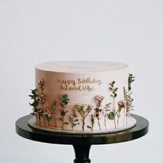 a pink birthday cake decorated with flowers on a black stand and happy birthday written in gold lettering