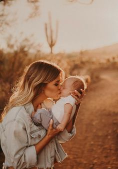 a woman holding a baby in her arms and kissing it's face with the sun behind her