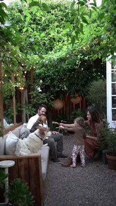 two people and a dog sitting on a couch in a garden area with potted plants