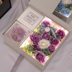 two white and purple roses in a box on a table with other flowers around it