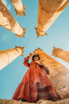 a woman in a red polka dot dress and hat standing between two palm trees with blue sky behind her