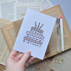 a hand holding up a card with a birthday cake on it next to some books