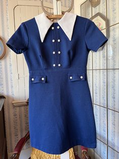 This cute dress comes from Wendy Lane, Montreal. It's made from fortrel in dark royal blue with a pointy wide fortrel collar and little white buttons down the front and on the faux pockets. It fastens with a long zipper in the back. The measurements, taken with the dress lying flat, are: shoulder to shoulder, 14 inches; armpit to armpit, 16 inches; sleeves, 7 inches; waist, 13 inches; length, 30 inches; bottom edge, 21 inches. In good condition. Preppy Fitted Mini Dress, Fitted Retro Dress With Doll Collar, Retro Fitted Dress With Doll Collar, Fitted Blue Dress With Doll Collar, Preppy Fitted Collared Dress, Preppy Fitted Knee-length Dresses, Vintage Blue Dress With Doll Collar, Blue Fitted Vintage Dress With Short Sleeves, Blue Short Sleeve Dresses With Buttons