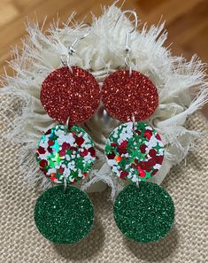 three pairs of christmas themed earrings on top of a pile of white feathers with red and green confetti