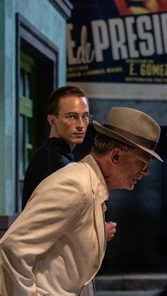 two men are walking down the street in front of a building and one is wearing a hat