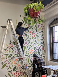 two people on ladders painting a wall with flowers and plants hanging from the ceiling