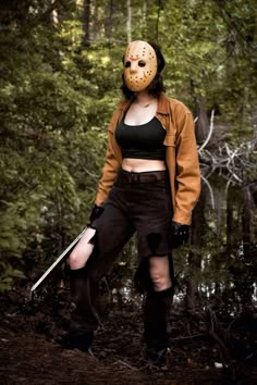 a woman wearing a mask and holding a knife in the woods with trees behind her