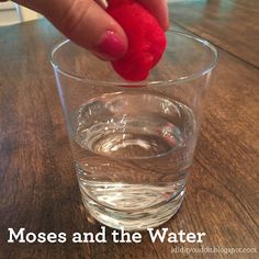 a hand holding a red object over a glass filled with water on top of a wooden table