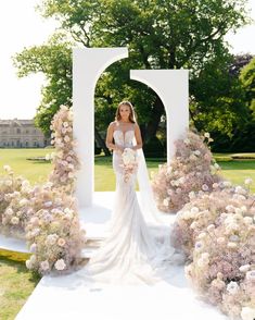 a woman standing in front of a white arch with flowers on the ground and bushes behind her