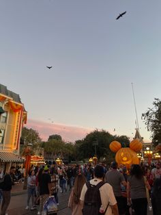 a crowd of people walking down a street next to tall buildings at dusk with flying birds overhead