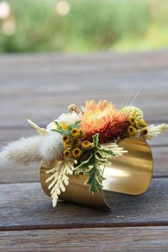 an arrangement of flowers in a gold vase on a wooden table with grass and feathers