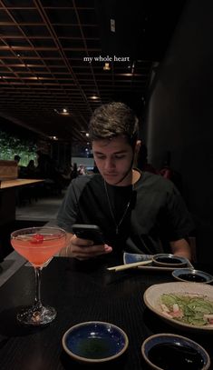 a young man sitting at a table looking at his cell phone while holding a drink