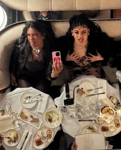 three women sitting at a table with food and drinks in front of them, taking a selfie