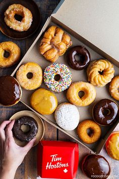 a box filled with lots of donuts on top of a wooden table next to a person's hand