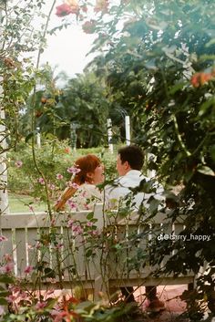 a man and woman sitting on a bench in the middle of a garden with pink flowers