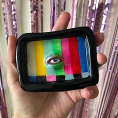 a hand holding up a small television with an eye on it's screen in front of some pink and purple sequins