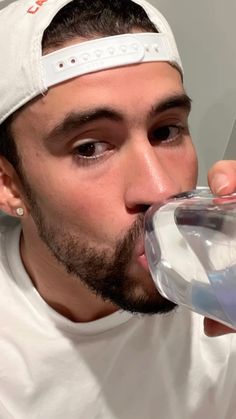 a man drinking water from a glass in front of his face and wearing a white hat