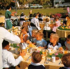 a group of children sitting at a table eating food and drinking orange juices while an adult stands in front of them