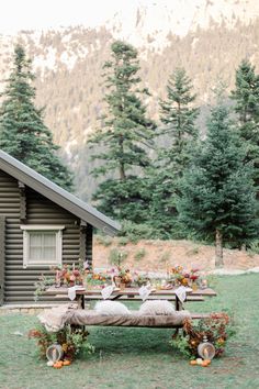 a picnic table set up in front of a log cabin with flowers and greenery