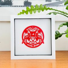 a red paper cut in a white frame on a wooden table next to a plant