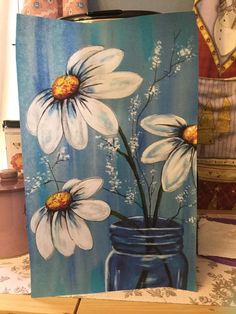 a painting of white daisies in a blue mason jar on a table next to other items