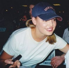 a woman sitting at a table with a microphone in her hand and wearing a baseball cap