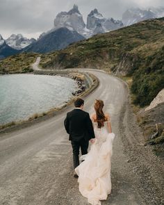 a bride and groom walking down the road in front of some mountains with snow capped peaks