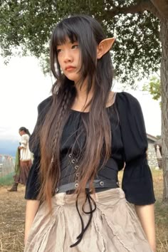 a woman with long hair wearing a skirt and black top standing in front of a tree