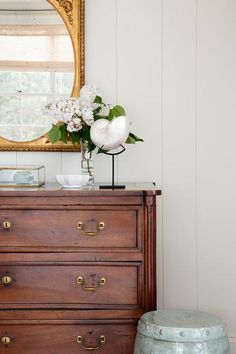 a dresser with flowers on top and a mirror in the corner above it next to a stool