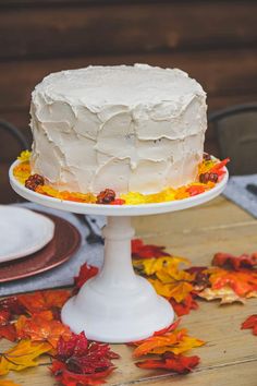 a white frosted cake sitting on top of a wooden table next to autumn leaves