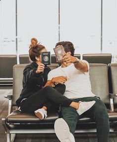 a man and woman sitting on a bench in an airport holding up their phones to take pictures