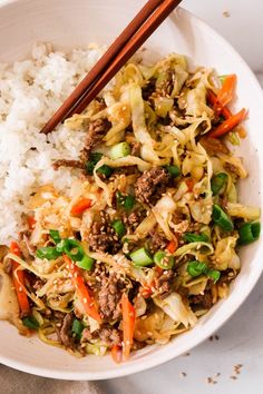 a white bowl filled with rice, meat and veggies next to chopsticks