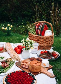 an outdoor picnic with food and drinks on the grass in front of some bushes, flowers and trees