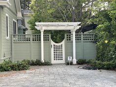 a white gate in front of a green house with trees and bushes around the entrance