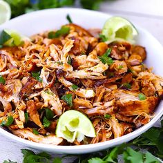 a white bowl filled with shredded chicken and garnished with cilantro, limes, and avocado